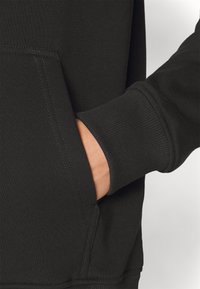 Felpa nera con tasca frontale, polsini a coste e cuciture laterali. Texture morbida con dettagli di cuciture visibili.