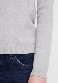 Harma neulepaita, jossa on ribbireunat, tekstuuri kangas, yhdistettynä tummansinisiin farkkuihin. Lähikuva hihasta ja helma yksityiskohdista.