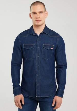 Man met kort haar, gekleed in een donkerblauw denim overhemd met twee borstzakken en bijpassende blauwe spijkerbroek, staand tegen een effen achtergrond.