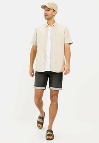 Beige Kurzarmhemd mit Knöpfen, darunter ein weißes T-Shirt, dunkle Denim-Shorts mit Umschlägen und schwarze Sandalen mit zwei Schnallen.