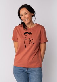 Rustfarbene Baumwoll-T-Shirt mit einem schwarz gezeichneten Profil einer Frau mit einem Dutt, akzentuiert durch Sonnen- und Mondmotive in einem minimalistischen Stil.