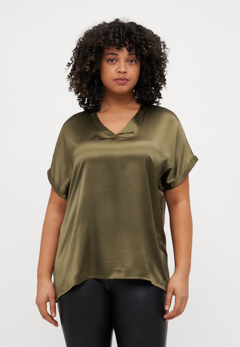 Blusa in satin verde oliva con scollatura a V e maniche corte arrotolate; vestibilità morbida con un orlo posteriore leggermente più lungo. Texture liscia e design elegante.
