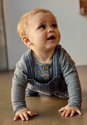 Jonge peuter met krullend blond haar die over de vloer kruipt, gekleed in een grijs geribd shirt met lange mouwen en een blauw gestreepte tuinbroek.