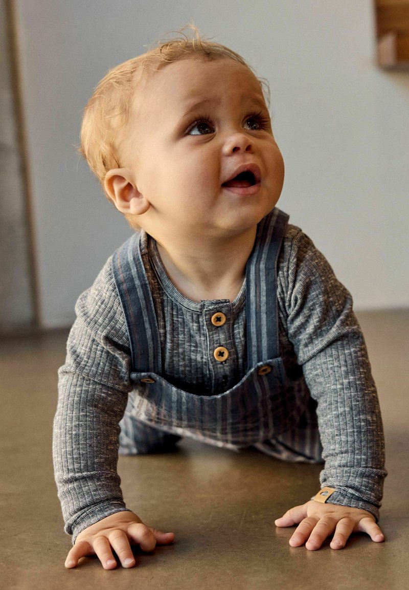 Jeune tout-petit aux cheveux blonds bouclés rampant sur un sol, portant un t-shirt gris côtelé à manches longues et une salopette rayée bleue.