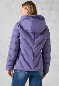 Lila puffjacka med huva, prydd med ett chevronmönster, ribbad textur och elastiska muddar, i kombination med blå denimjeans.