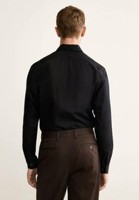 Maglietta nera a maniche lunghe con colletto standard, realizzata in tessuto liscio, abbinata a pantaloni marroni con tasche posteriori e dettagli con bottoni.