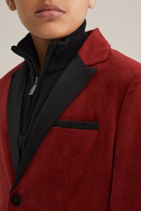 Blazer en velours côtelé rouge avec un revers en satin noir à cran et des détails de poche, superposé sur un pull à fermeture éclair noir. Tissu texturé et coupe ajustée.