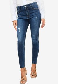 Vysoké modré džíny skinny s potrhanými detaily na kolenou a třásněmi na lemech, v kombinaci s páskovými metalickými sandály.