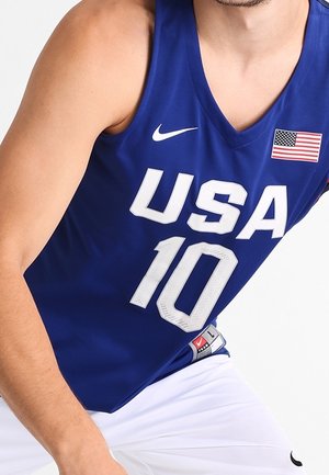 Sportowiec ubrany w niebieską koszulkę do koszykówki USA z numerem 10 i naszywką z flagą Ameryki, w połączeniu z białymi szortami.