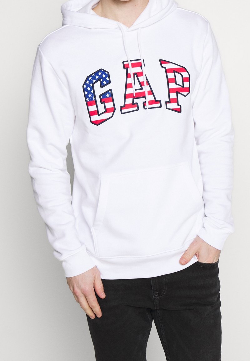 Sweat à capuche blanc avec une poche avant et un cordon de serrage à la capuche. Présente le mot « GAP » en lettres majuscules avec des motifs du drapeau américain en bleu et rouge.