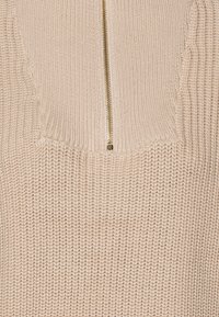 Beige stickad tröja med texturerad yta och dragkedja framtill. Har en vid halsringning och ribbade kanter för struktur.