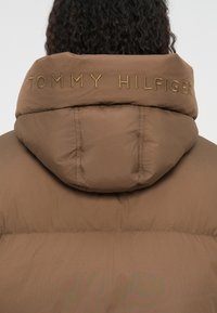 Brun pufferjakke med hætte, prydet med guldbroderet "TOMMY HILFIGER" på ryggen, glat tekstur og syede detaljer.