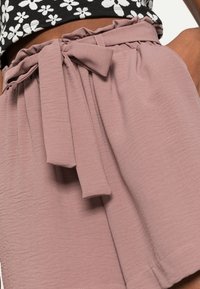 Růžové kraťasy s volány a texturovanou tkaninou, s širokým pasem a mašlí, ukazující volný střih a čistý lem.