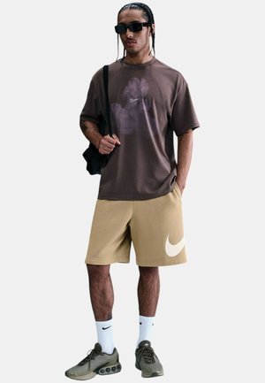 T-shirt grafica marrone con logo, pantaloni corti beige con un lato a swoosh, calzini bianchi e sneakers verde oliva. Inclusi occhiali da sole neri e zaino.