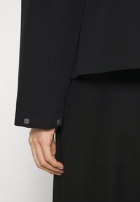 Black tailored jacket featuring a smooth fabric, sleek design, and a cuff with button accents. Simple and classic cut for versatility.