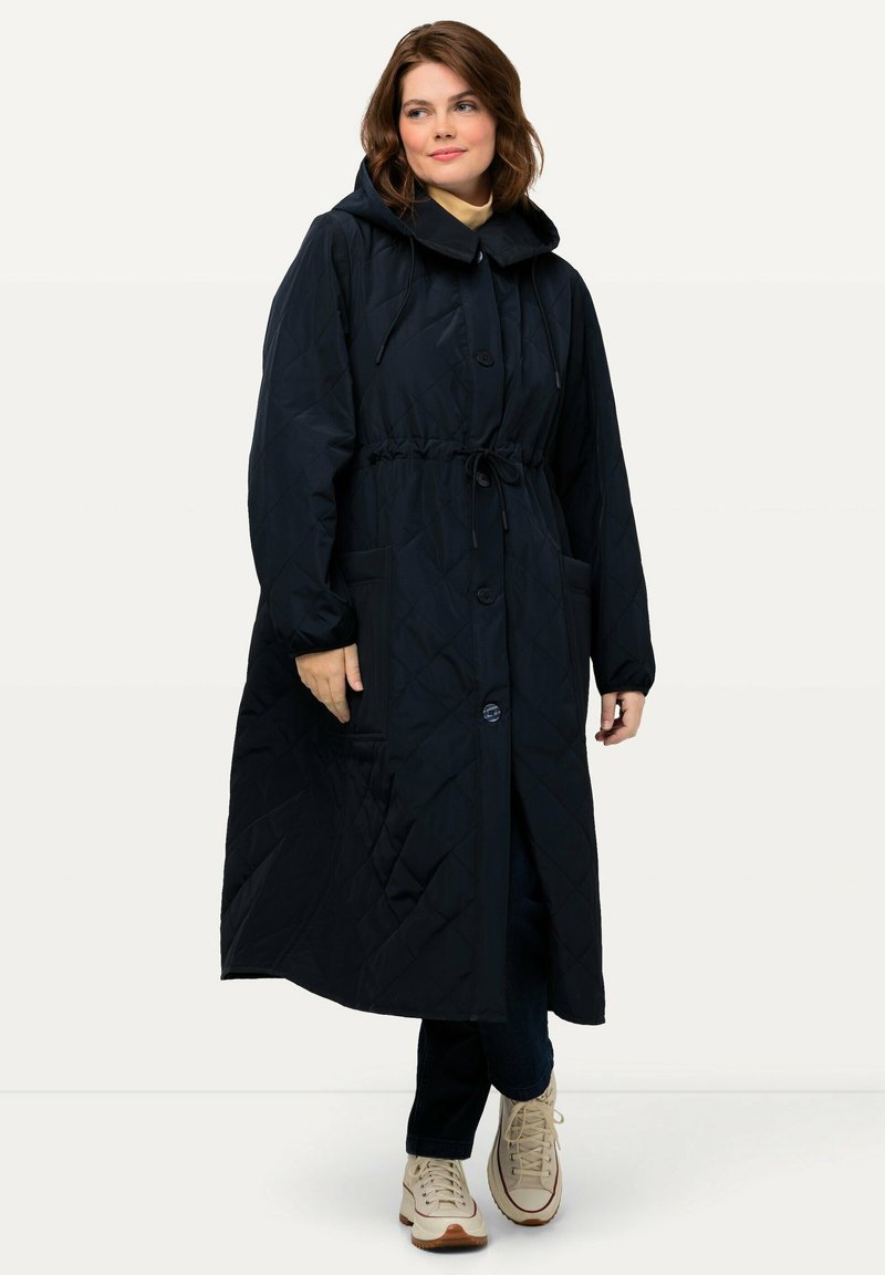 Quiltad marinblå lång kappa med huva, som har dragsko i midjan, knappstängning och sidofickor. Mjukt tyg med en släta yta.