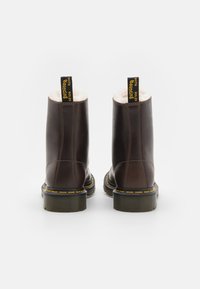 Bottines en cuir marron avec doublure blanche en peluche, couture jaune et semelles en caoutchouc noir. Dotées d'une languette noire à l'arrière.