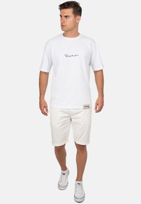 T-shirt blanche à manches courtes avec logo noir, associé à des shorts blancs retroussés et des baskets blanches avec des accents rouges.