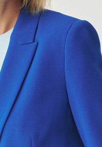 Královský modrý sako z texturované látky, s výrazným klopovým límcem a jednou přední kapsou, zobrazeno od ramene dolů.