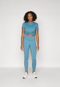 Turkis aktivtøy som har en knyttet crop topp og høyt midjeleggings med et subtilt mønster, sammen med hvite joggesko.