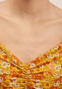 Top floral avec un décolleté en V de couleur jaune, orné de fleurs orange, rouges et blanches, avec une texture froncée au centre.