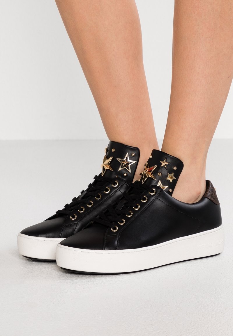 Baskets basses noires avec semelles blanches, clous dorés en forme d’étoile sur la languette et œillets dorés assortis, portées pieds nus.