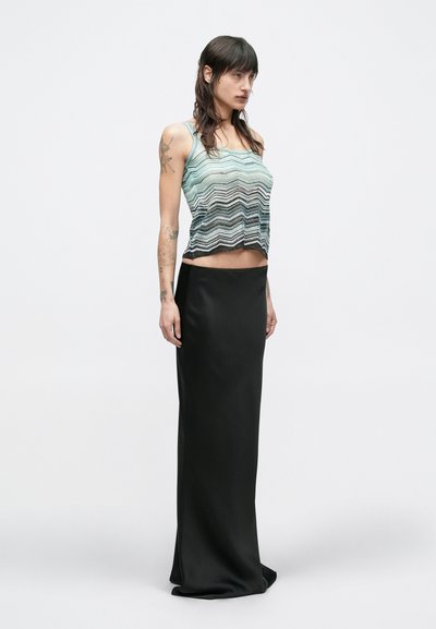 Γυναίκα με μακριά σκουρόχρωμα μαλλιά και τατουάζ, φορεί ριγωτό τοπ χωρίς μανίκια και μακριά μαύρη φούστα, στέκεται σε απλό λευκό φόντο.