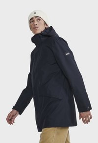 Marineblauwe waterdichte jas met een capuchon, voorzien van een voorzak, verstelbare manchetten en een logo-opdruk op de mouw. Draag met beige broek.