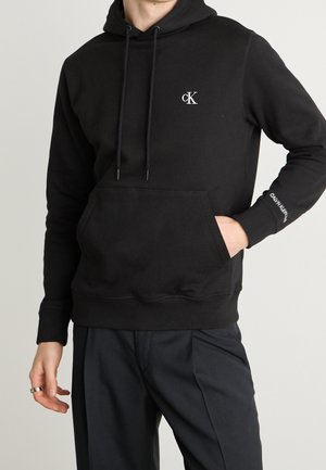 Czarna bluza z kapturem z przednią kieszenią, białe logo "cK" na piersi, napis "Calvin Klein" na rękawie, noszona przez osobę z ręką w kieszeni.