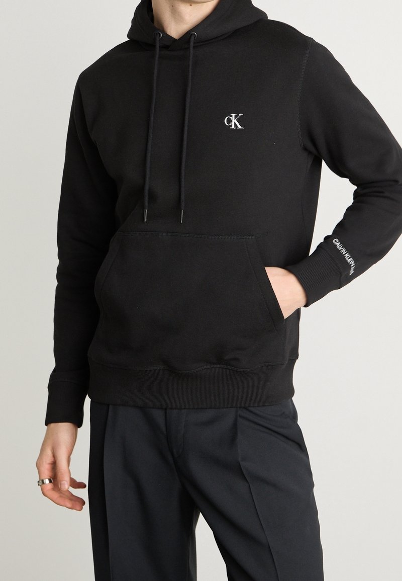 Sweat à capuche noir avec poche avant, logo blanc "cK" sur la poitrine, texte "Calvin Klein" sur la manche, porté par une personne avec la main dans la poche.