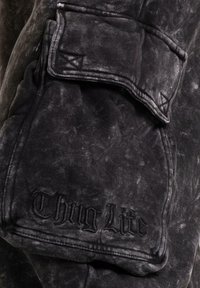 Svart denimstoff med slitt finish, med en klafflomme og brodert "Thug Life"-tekst. Strukturert og robust design.