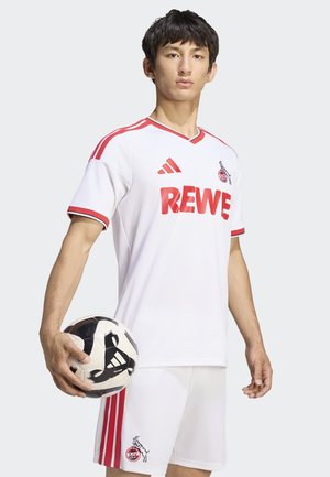 Junger Mann in weiß-rotem Fußballtrikot hält einen schwarz-weißen Adidas Fußball vor einem einfarbigen hellen Hintergrund.