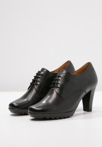 Chaussures à talons hauts en cuir noir avec une texture lisse, un design à lacets et une semelle rainurée. Dotées d'un bout pointu et d'un talon en bois.