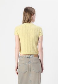 Kratka, prilegajoča rumena pletena majica s teksturiranim pletenim vzorcem, kratkimi rokavi in skrajšanim robom, nošena z visokim pasom svetlo modre jeans krila.