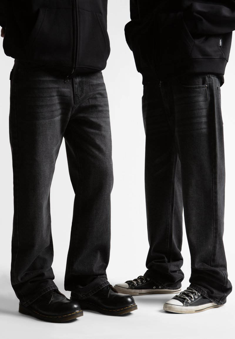 Δύο άτομα φορούν μαύρα φούτερ με κουκούλα, σκούρα γκρι τζιν· το ένα με μαύρα δερμάτινα παπούτσια, το άλλο με μαύρα και άσπρα αθλητικά παπούτσια, στέκονται δίπλα δίπλα.