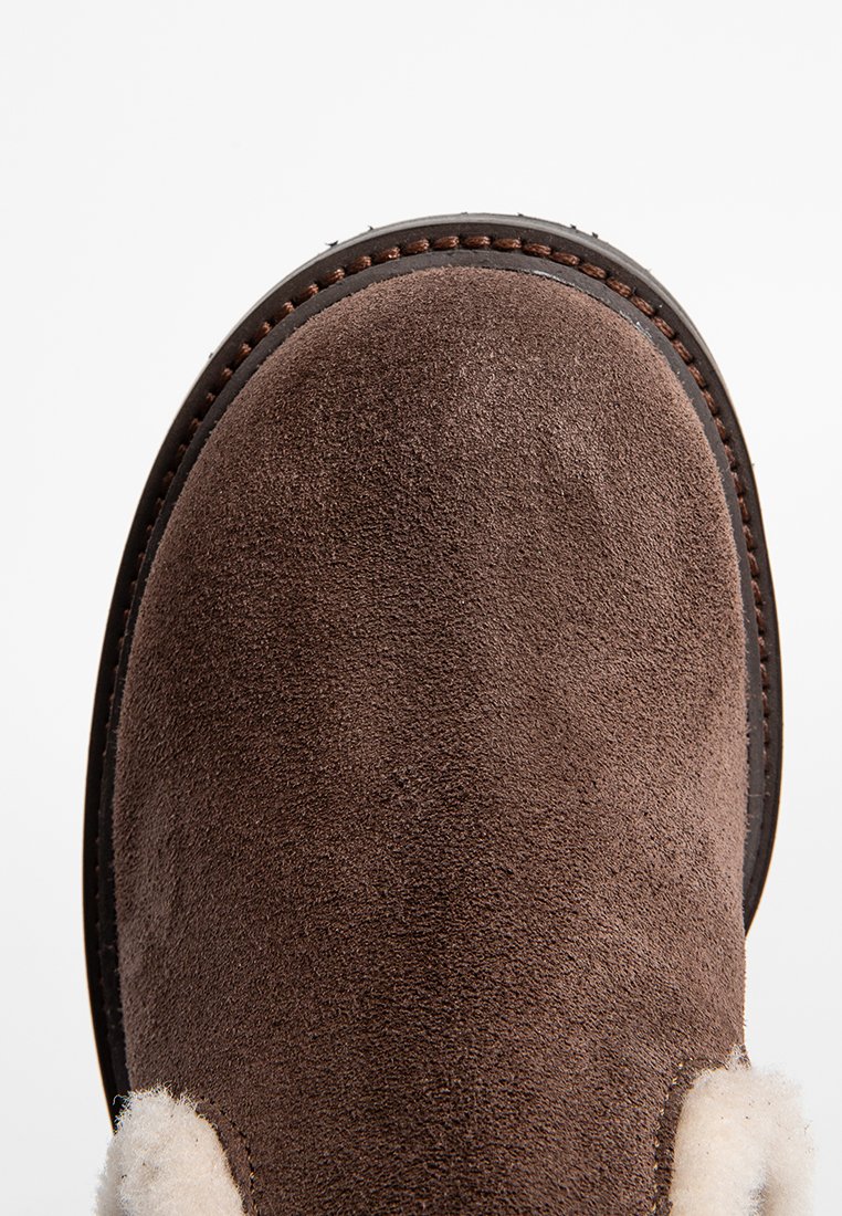 Bruine suède enkellaars met een ronde neus, zachte witte schapenvachtkraag en contrasterende donkerbruine stiksels langs de rand.