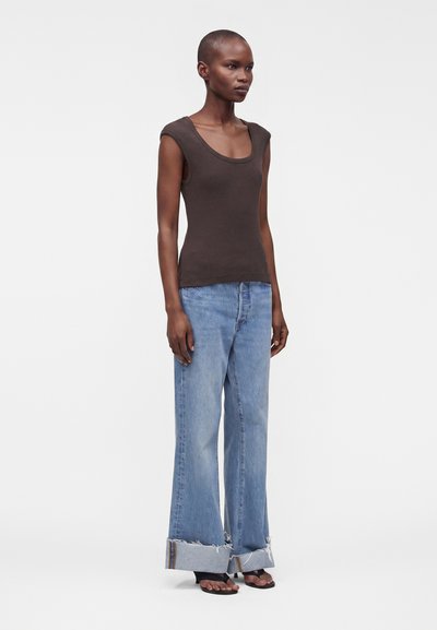 Femme debout portant un haut sans manches marron foncé, un jean large bleu clair aux ourlets effilochés et des sandales noires à bouts ouverts sur un fond uni.