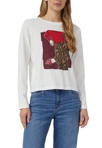 Biała bluzka z długim rękawem z graficznym wzorem przedstawiającym osobę w spodniach w czerwono-brązowy wzór. Połączona z niebieskimi dżinsami i wzorzystymi butami.