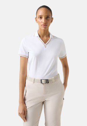 Vrouw draagt een witte poloshirt met korte mouwen en beige biezen, beige broek en een geweven beige riem, staand tegen een effen grijze achtergrond.