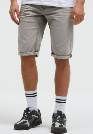 Unterkörper einer Person, die hochgekrempelte hellgraue Shorts, weiße Socken mit schwarzen Streifen und schwarz-graue Sportschuhe mit blauen Akzenten trägt.