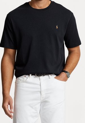 Man draagt een zwart T-shirt met korte mouwen met een klein geborduurd logo en witte broek, met één hand in de zak en een horloge om.