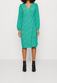 Grünes Wickelkleid mit V-Ausschnitt, langen Ärmeln und einem schwarzen abstrakten Muster. Kombiniert mit schwarzen Knöchelstiefeln. Glatter Stofftextur.