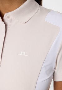 Jasnobeżowa koszulka polo z białymi akcentami na ramieniu i rękawie, z małym białym stylizowanym logo na klatce piersiowej.