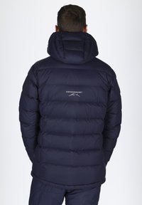 Mörkblå puffjacka med huva, prydd med ett quiltat mönster och logotyp på baksidan. Slät textur och lätt design.