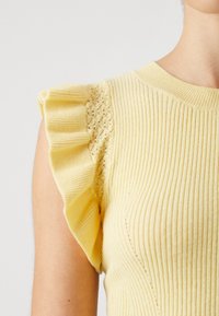 Bezramionny żółty pleciony top z koronkowymi detalami i falbanami na ramieniu, noszony przez osobę na jednolitym tle.