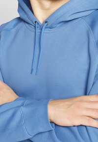 Sudadera de algodón azul con una textura suave, capucha ajustable con cordones y puños de canalé. Ofrece un ajuste relajado y costuras limpias.
