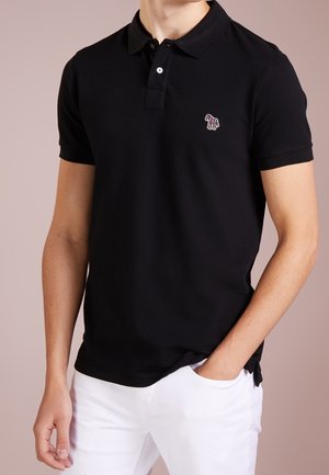 Man draagt een zwart poloshirt met korte mouwen met een kleurrijk geborduurd zebra-embleem en witte broek, hand in zak, neutrale achtergrond.