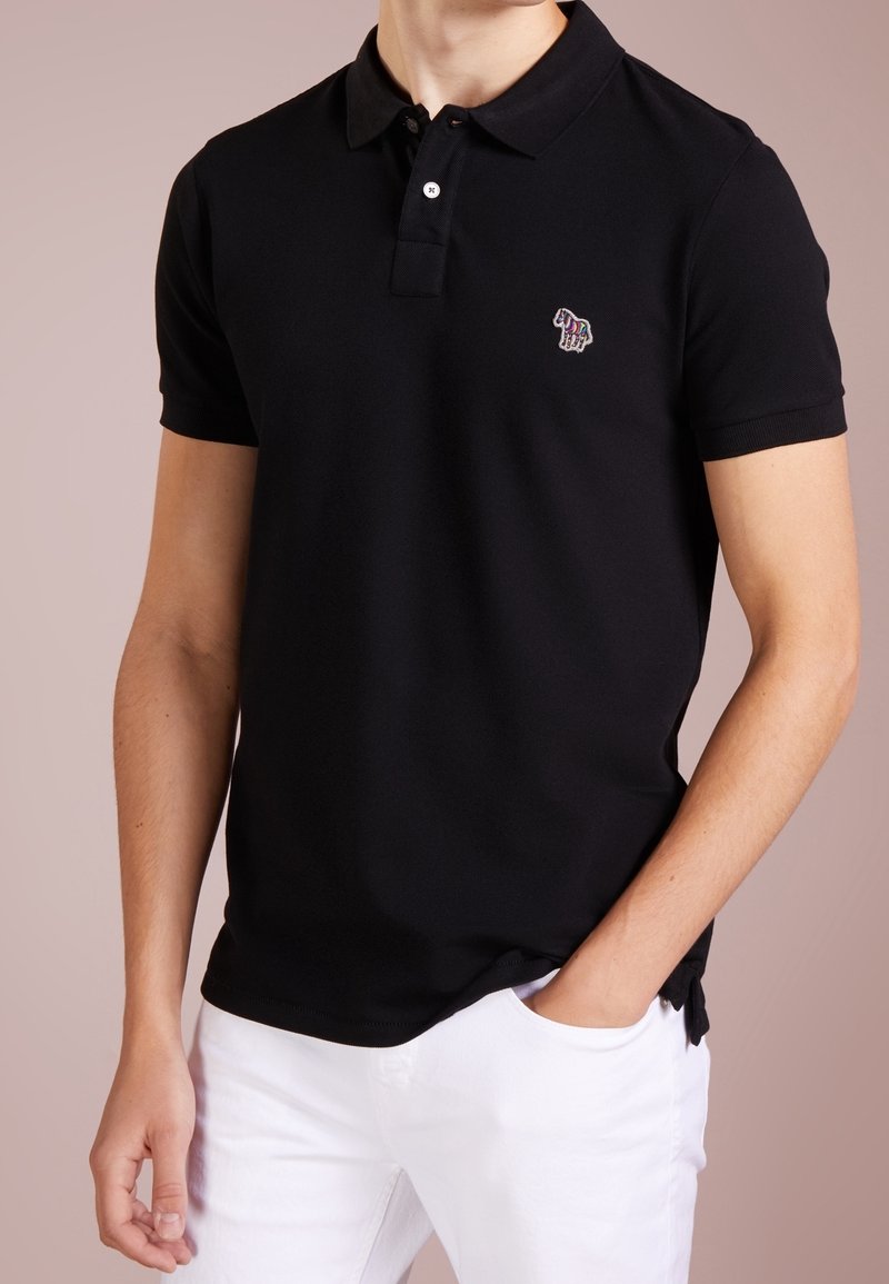 Homme portant un polo noir à manches courtes avec un emblème zèbre brodé coloré et un pantalon blanc, main dans la poche, fond neutre.