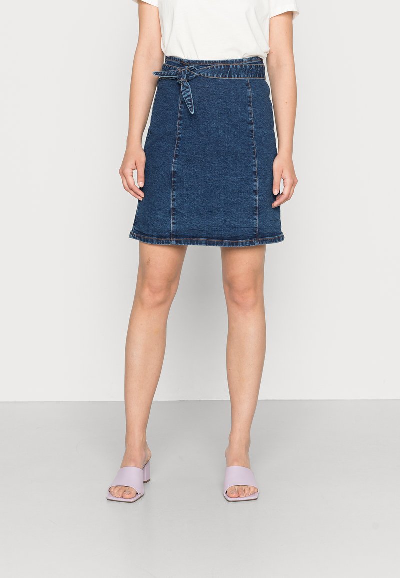 Saia de ganga, azul escuro, acima do joelho, com um cinto atado na cintura e costuras em contraste. Combinada com sandálias de salto bloco em lavanda.