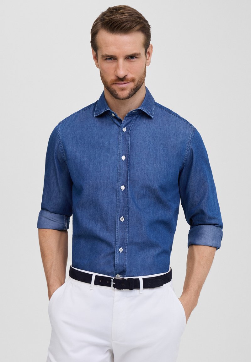Camicia di denim abbottonata di colore blu scuro, con colletto, maniche arrotolate e bottoni bianchi. Abbinata a pantaloni bianchi e una cintura scura.